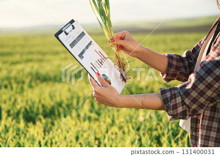Side view. With notepad and plant. Young woman is on the beautiful agricultural field at daytime Side view. With notepad and plant. Young woman is on the beautiful agricultural field at daytime 131400031