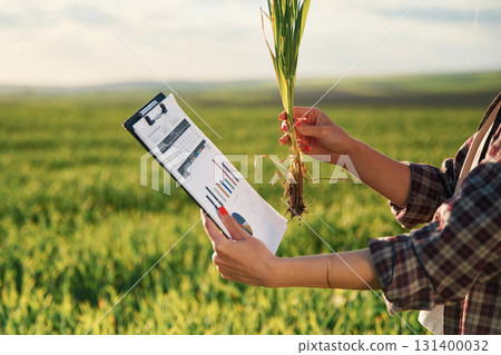 Side view. With notepad and plant. Young woman is on the beautiful agricultural field at daytime Side view. With notepad and plant. Young woman is on the beautiful agricultural field at daytime 131400032
