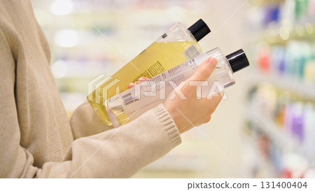 Close up of woman comparing two shampoo bottles in a beauty store, reading labels and ingredients. Female shopper choosing haircare product. Concept of conscious choice and healthy lifestyle. Close up of woman comparing two shampoo bottles in a beauty store, reading labels and ingredients. Female shopper choosing haircare product. Concept of conscious choice and healthy lifestyle. 131400404