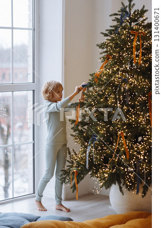 Child decorating Christmas tree with ribbons in cozy room with white wall and large window 131400671