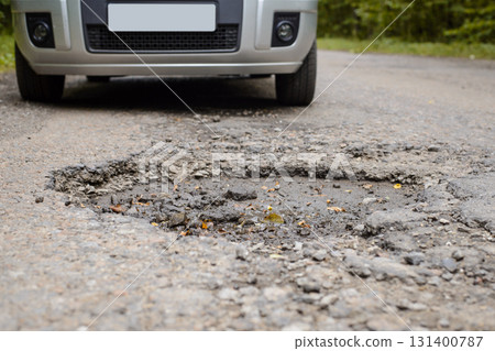 Silver car stopped in front of big crack in asphalt surface. Rural road in terrible condition and needs repairing, enormous pothole on a highway in countryside 131400787