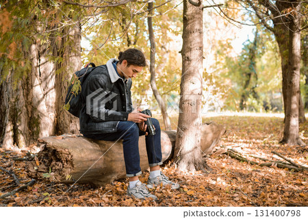 Young nature photographer sitting on log in forest, looking pictures after successful animals and nature photoshoot, satisfied with results Young nature photographer sitting on log in forest, looking pictures after successful animals and nature photoshoot, satisfied with results 131400798