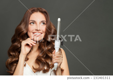 Glorious woman with long wavy hairstyle using hair curling tongs iron, indoor portrait Glorious woman with long wavy hairstyle using hair curling tongs iron, indoor portrait 131401311