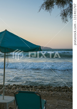 Beach scene at sunset with turquoise umbrella and sun loungers, small table between loungers, sea with small waves 131401707