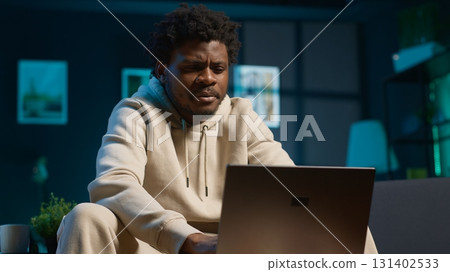Young male freelancer managing schedules and notes on his laptop while seated in a contemporary living room. Inspiring man looks at pc for a productive teleworking session. Camera A. Young male freelancer managing schedules and notes on his laptop while seated in a contemporary living room. Inspiring man looks at pc for a productive teleworking session. Camera A. 131402533