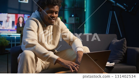 Confident young man solving daily tasks on a freelance platform at home, answering emails and multitasking. Person staying connected and enjoying the flexibility of working remotely. Camera B. 131402534