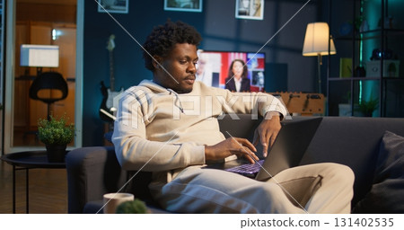 Black man does his research by reading articles and taking notes at home, collecting information for a new project. Freelancer multitasking and studying files with data driven papers. Camera B. 131402535