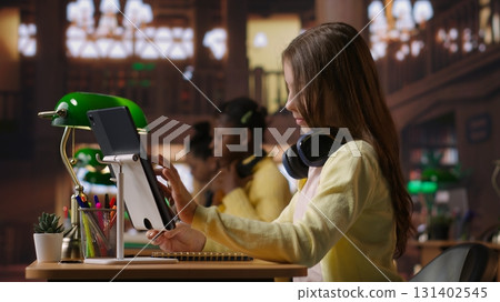 Preteen student using a tablet to complete her class notes in the library, uses he digital academic database to consult learning materials. Preparing diligently to pass her exams. Camera B. Preteen student using a tablet to complete her class notes in the library, uses he digital academic database to consult learning materials. Preparing diligently to pass her exams. Camera B. 131402545