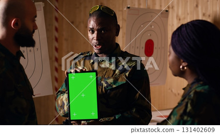 Soldiers at shooting range do training designed to increase safety and survivability in combat, watching instructional videos on green screen chroma key tablet device presented by commander, camera B 131402609