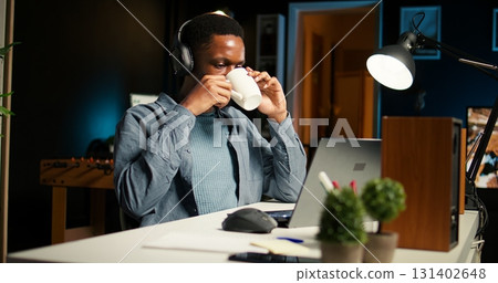 African american specialist closing the monthly balance with a show on headset, working remotely while he listens to his favorite podcast. Managing expenses and insurances from home. Camera A. 131402648