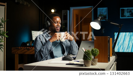 Young casual guy listening to a podcast while he works on freelance job at home, enjoys multitasking with headphones. Man solving tasks and paying attention to webinar discussion. Camera A. Young casual guy listening to a podcast while he works on freelance job at home, enjoys multitasking with headphones. Man solving tasks and paying attention to webinar discussion. Camera A. 131402663