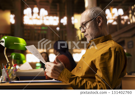 Elderly man with a serious expression studies at a college library desk, engaged in academic research and reading textbooks to complete his degree and enrich his understanding of literature. Elderly man with a serious expression studies at a college library desk, engaged in academic research and reading textbooks to complete his degree and enrich his understanding of literature. 131402760