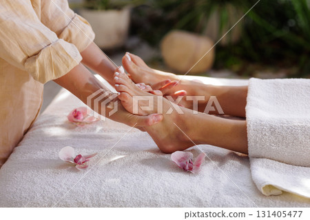Therapist performs soothing foot reflexology on a spa table outdoors with orchids and soft daylight, ideal for wellness and relaxation themes. Therapist performs soothing foot reflexology on a spa table outdoors with orchids and soft daylight, ideal for wellness and relaxation themes. 131405477