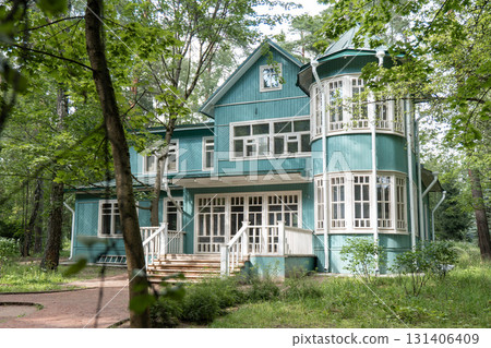 A historic wooden house in Peredelkino is painted in teal with white window frames, surrounded by tall trees and summer greenery 131406409