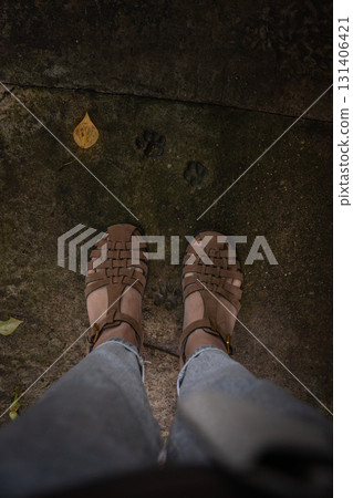 Brown sandals and light jeans are seen on an old concrete surface with visible animal paw prints and a yellow leaf Brown sandals and light jeans are seen on an old concrete surface with visible animal paw prints and a yellow leaf 131406421