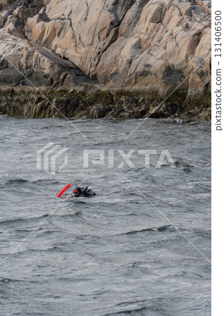 A scuba diver in black gear with a red buoy floats near rocky coastal cliffs under cloudy skies A scuba diver in black gear with a red buoy floats near rocky coastal cliffs under cloudy skies 131406500
