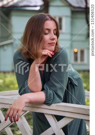 A woman in a green dress leans on a white wooden fence, looking confidently at the camera with a turquoise house in the background 131406520