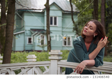 A woman in a green dress leans on a white wooden fence, gently touching her hair with closed eyes against the backdrop of a turquoise country house 131406540