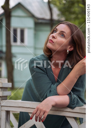 A woman in a dress leans thoughtfully on a white wooden fence, gazing upward with an expressive look against the backdrop of a turquoise country house 131406543