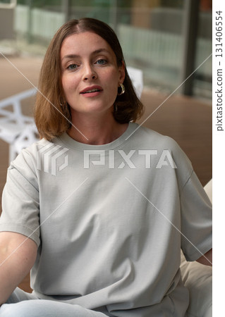 A woman in a light gray t-shirt and hoop earrings looks into the camera with a gentle smile, seated on a modern terrace 131406554