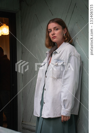 A woman in a light denim jacket and green dress standing against a rustic wooden wall, looking directly at the camera with a serious expression 131406555