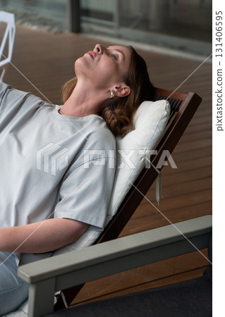 A woman in a light gray t-shirt reclines on a cushioned wooden lounge chair, gazing upward in a calm, reflective mood A woman in a light gray t-shirt reclines on a cushioned wooden lounge chair, gazing upward in a calm, reflective mood 131406595
