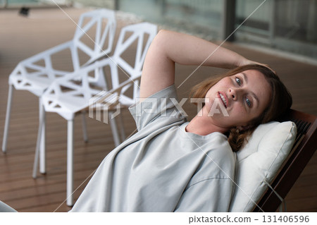 A relaxed woman in a light gray t-shirt lounges on a cushioned wooden chair, smiling softly toward the camera 131406596