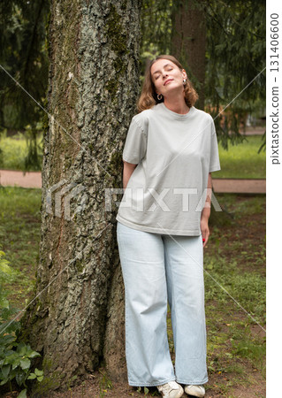 Woman in a gray t-shirt and light jeans leans against a tree with closed eyes, enjoying the moment in a green park 131406600