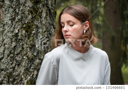 Woman in a light gray t-shirt leans against a tree, looking down thoughtfully with a calm expression Woman in a light gray t-shirt leans against a tree, looking down thoughtfully with a calm expression 131406601