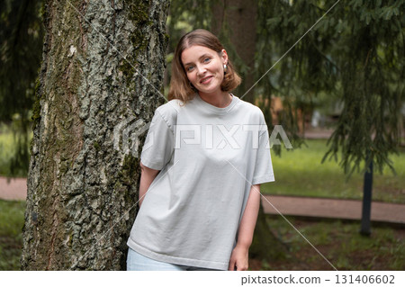 Smiling woman in a light gray t-shirt leans casually against a tree in a green park in Peredelkino 131406602