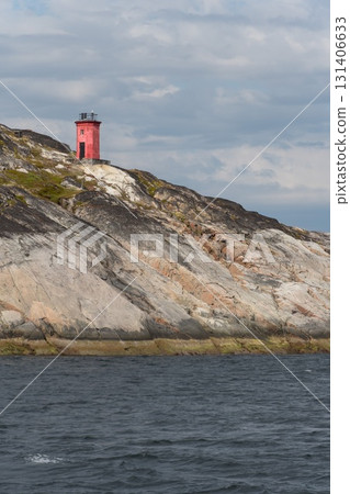 A small red lighthouse stands on rugged rocky cliffs above dark blue water under cloudy skies A small red lighthouse stands on rugged rocky cliffs above dark blue water under cloudy skies 131406633