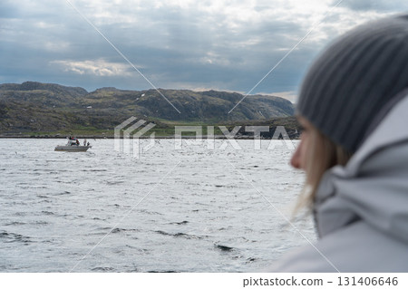 A person in a white jacket and gray knitted hat looks at small boats on the Barents Sea under a cloudy sky A person in a white jacket and gray knitted hat looks at small boats on the Barents Sea under a cloudy sky 131406646