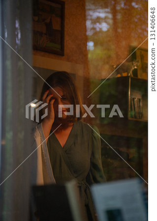 A young woman in a green dress is seen through a window, softly touching her face in a cozy vintage-style room with warm lighting. Peredelkino A young woman in a green dress is seen through a window, softly touching her face in a cozy vintage-style room with warm lighting. Peredelkino 131406648