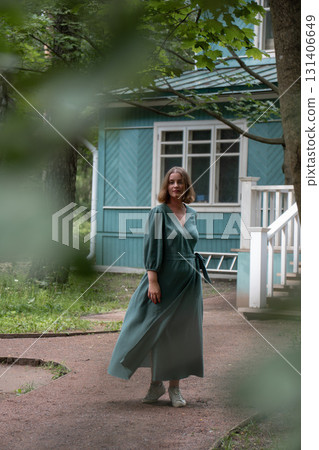 A woman in a flowing green dress spins gracefully on a garden path in front of a wooden house 131406649