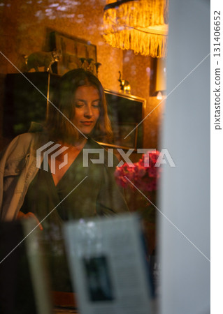 A woman in a green dress is seen through a window in a cozy vintage room with warm lighting and a bouquet of flowers. Peredelkino 131406652
