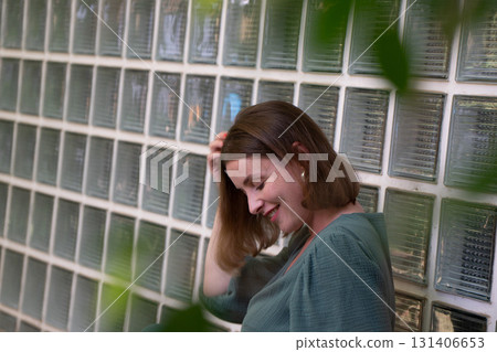 Woman in a green dress sits near a wall of glass blocks, smiling with her head tilted and hand in hair, surrounded by soft greenery 131406653