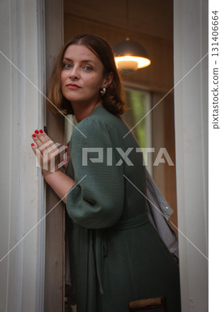 Woman in a green dress leans against a doorway, gazing softly at the camera with natural expression and warm light Woman in a green dress leans against a doorway, gazing softly at the camera with natural expression and warm light 131406664