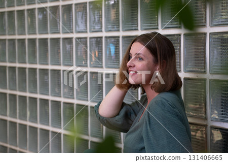 Woman in a green dress smiles while sitting against a wall of glass blocks, her hand resting near her hair Woman in a green dress smiles while sitting against a wall of glass blocks, her hand resting near her hair 131406665