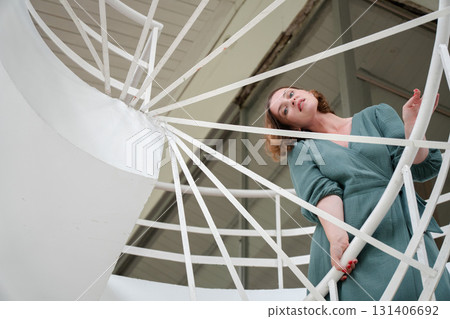 Woman in a green dress leans on a white spiral staircase, captured from a low angle with graphic architectural lines 131406692