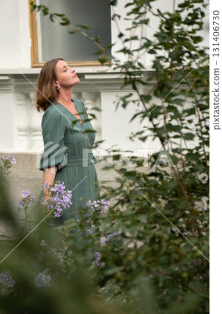 A woman in a green dress stands beside blooming purple flowers, her face raised peacefully toward the sky A woman in a green dress stands beside blooming purple flowers, her face raised peacefully toward the sky 131406730
