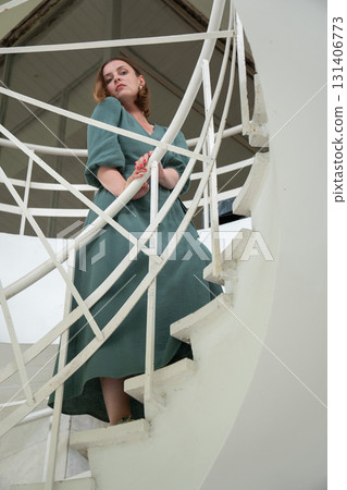 Woman in a green dress leans on a white spiral staircase, captured from a low angle with graphic architectural lines Woman in a green dress leans on a white spiral staircase, captured from a low angle with graphic architectural lines 131406773