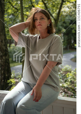 Young woman with short brown hair sits on a veranda in soft morning light, touching her hair thoughtfully 131406791