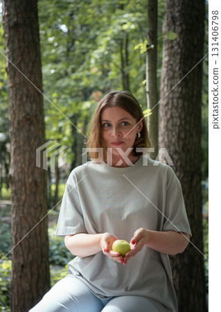Young woman in a light grey T-shirt tosses a green apple while smiling in a pine forest in Peredelkino, Russia 131406798