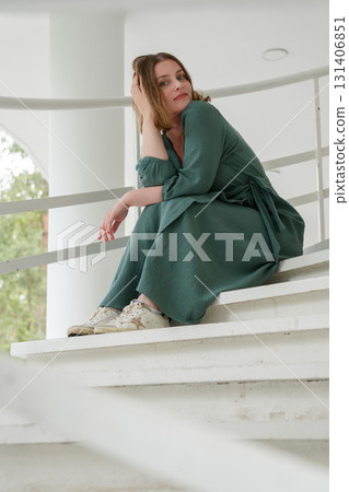 Woman in a green dress sits on white stairs in Peredelkino, leaning on the railing with a thoughtful, stylish pose 131406851