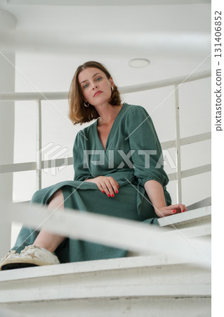 Woman in a green dress sits on white stairs in Peredelkino, leaning on the railing with a thoughtful, stylish pose 131406852