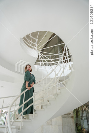 Woman in a green dress poses gracefully on a modern white spiral staircase in Peredelkino Woman in a green dress poses gracefully on a modern white spiral staircase in Peredelkino 131406854