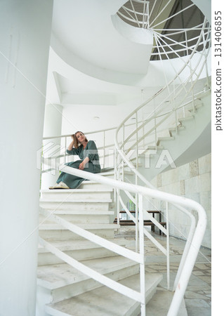 Woman in a green dress sits on a white spiral staircase in Peredelkino, leaning casually on the railing with a soft smile Woman in a green dress sits on a white spiral staircase in Peredelkino, leaning casually on the railing with a soft smile 131406855