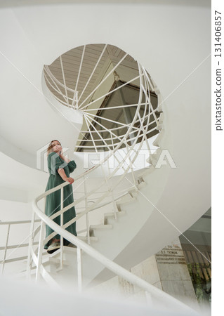 Woman in a green dress poses gracefully on a modern white spiral staircase in Peredelkino Woman in a green dress poses gracefully on a modern white spiral staircase in Peredelkino 131406857