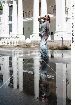 Woman in casual clothes stands near a neoclassical building, reflected in a large puddle on the pavement after rain 131406861