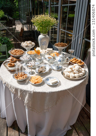 Outdoor vintage tea table with traditional Russian sweets, baranki, and chamomile bouquet in a porcelain vase, set against a wooden terrace 131406874